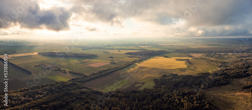 Wide panorama of flying high over countryside fields and forests. Dramatic sky during sunset, overcast sky with some places where sun rays come through thick clouds, drone point of view