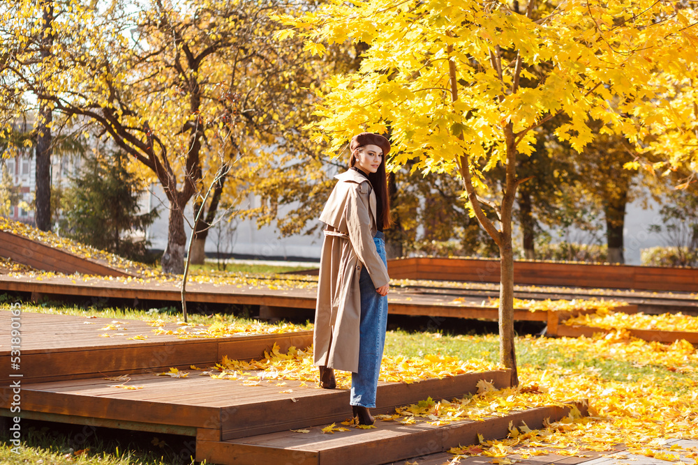 Stylish woman in beige trench and brown beret spending time outdoors in the park in autumn, yellow foliage all around. 