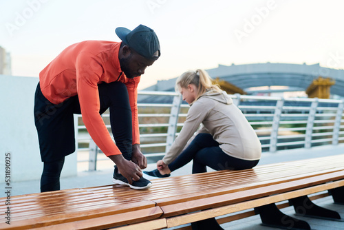 Young black man in activewear bending over leg bent in knee while tying shoelace of sneaker against sportswoman sitting on bench
