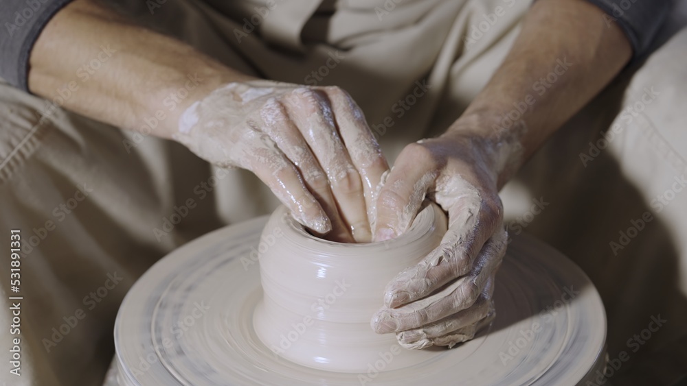 Wet male potter's hands sculpting soft clay and shaping the pot on potters wheel. Master gives shape to the pottery in a creative workshop. Creative masterclass from an artisan. Clay shaping close up.