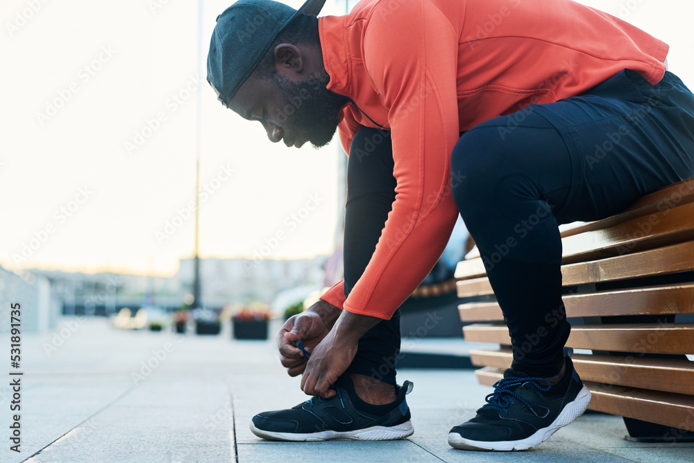 Side view of young African American runner in black leggins and cap and red sport jacket bending over sneaker while tying shoelace Side view of young African American runner in black leggins and cap and red sport jacket bending over sneaker while tying shoelace