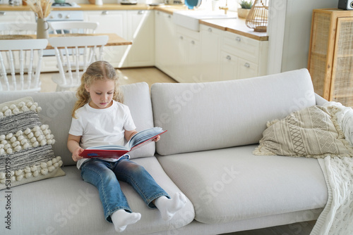 Small girl preschool child reads fairy tale stories holding paper book sitting on sofa alone at home