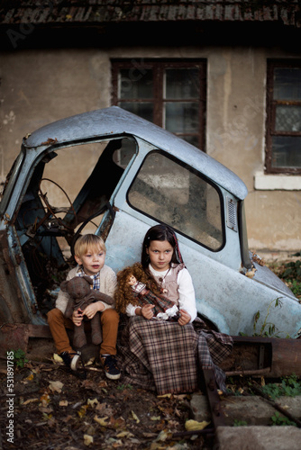 Biedne zaniedbane dzieci siedzą ze swoimi ulubionymi misiami i lalkami przy zepsutym starym samochodzie