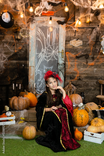 Cheerful child in witch costume sitting in decorated secrets room on wooden background, surround by web pumkins. Kids trick or treating. Beautiful brunette teenager girl celebrates Halloween carnival