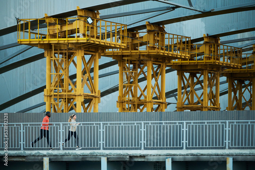 Row of huge architectural constructions standing along bridge with two runners jogging by riverside in the morning in urban environment