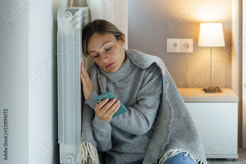 Young woman covered with warm blanket loses sense of living and motivation after seeing debts on phone screen. Female leaning on cold heating radiator scrolls phone looking for occupation to pay bills