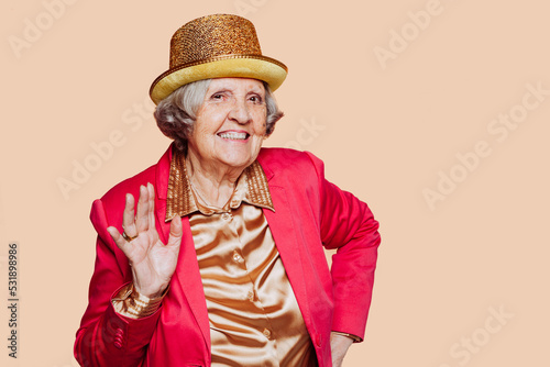 Active joyful stylish retired elder grandmother, wears hat, pink suit and formal golden shirt, posing at studio, has charming smile on face, isolated on beige wall, copy space area for advert.