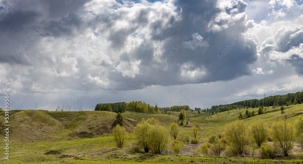 Spring landscape, dramatic sky over green hills before rain. The sun through the clouds illuminates the green meadows and ravines.