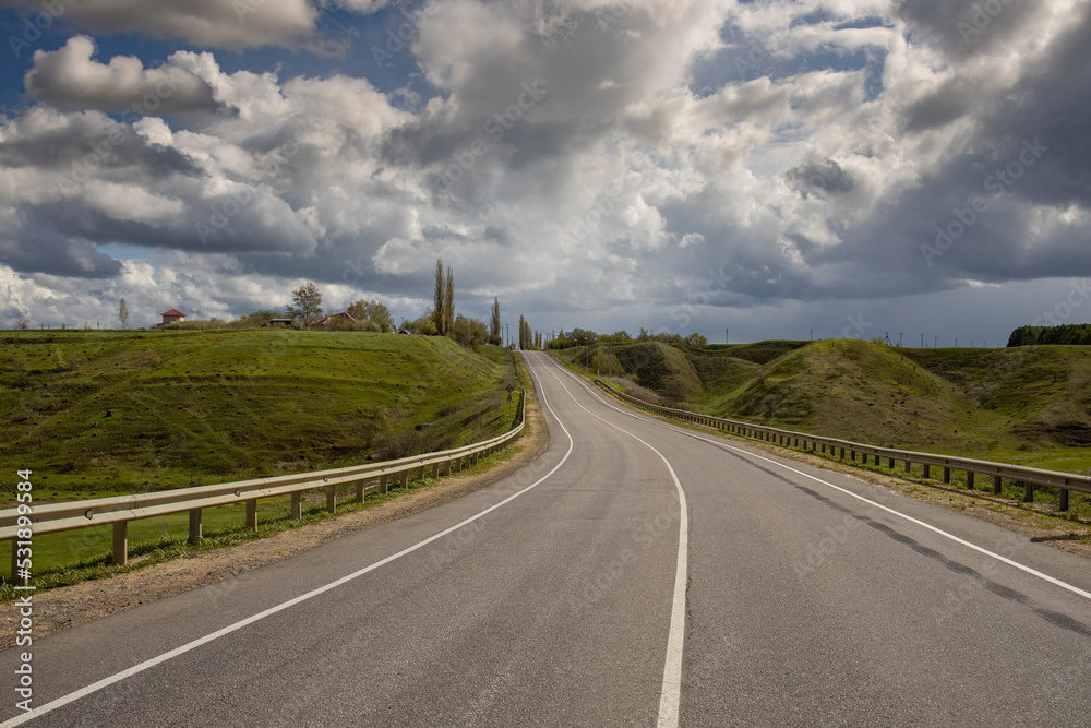 Osphalt road. Spring landscape, dramatic sky over green hills, before rain. The sun through the clouds illuminates the road and ravines. Osphalt road. Spring landscape, dramatic sky over green hills, before rain. The sun through the clouds illuminates the road and ravines.
