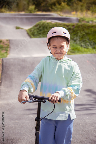  portrait of cute little caucasian school girl wear helmet enjoy having fun riding scooter on asphalted track.in street park outdoors on sunny day. Healthy sport children activities outsid