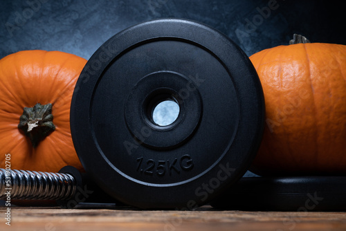 Fototapeta Naklejka Na Ścianę i Meble -  Heavy dumbbell barbell black weight plates with orange pumpkins. Healthy fitness lifestyle autumn fall composition for Halloween or Thanksgiving. Gym workout and sport training concept.