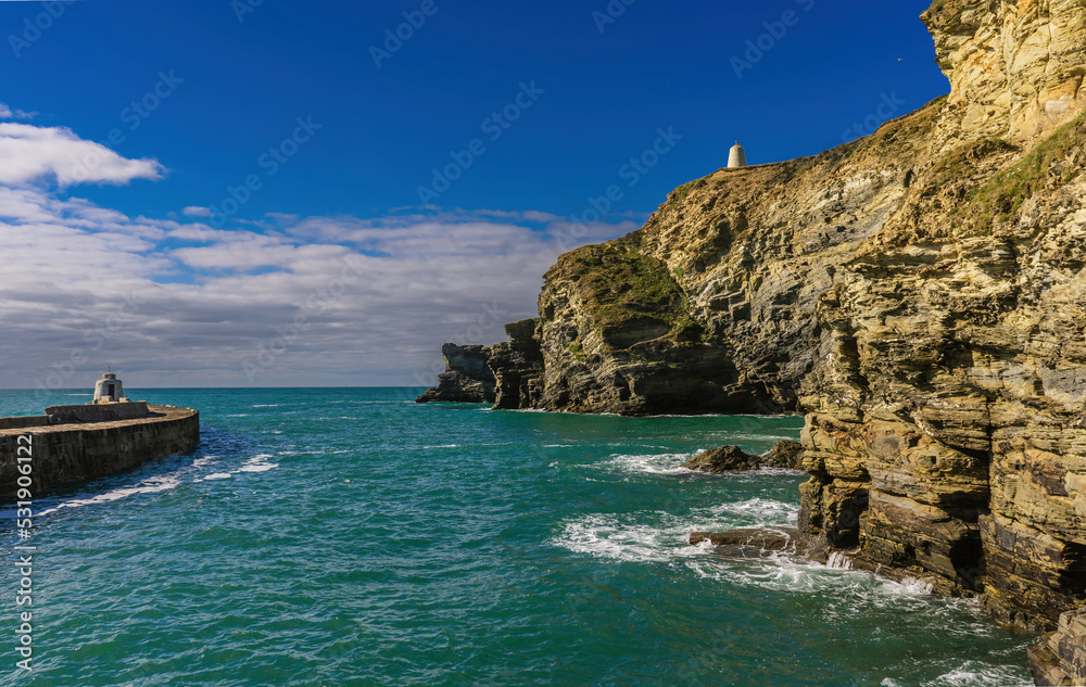 Water channel from the harbour in Portreath, Cornwall. On one side is the pier and on the other, cliffs, There is a lookout tower at the end of the pier and one high up on the cliffs.