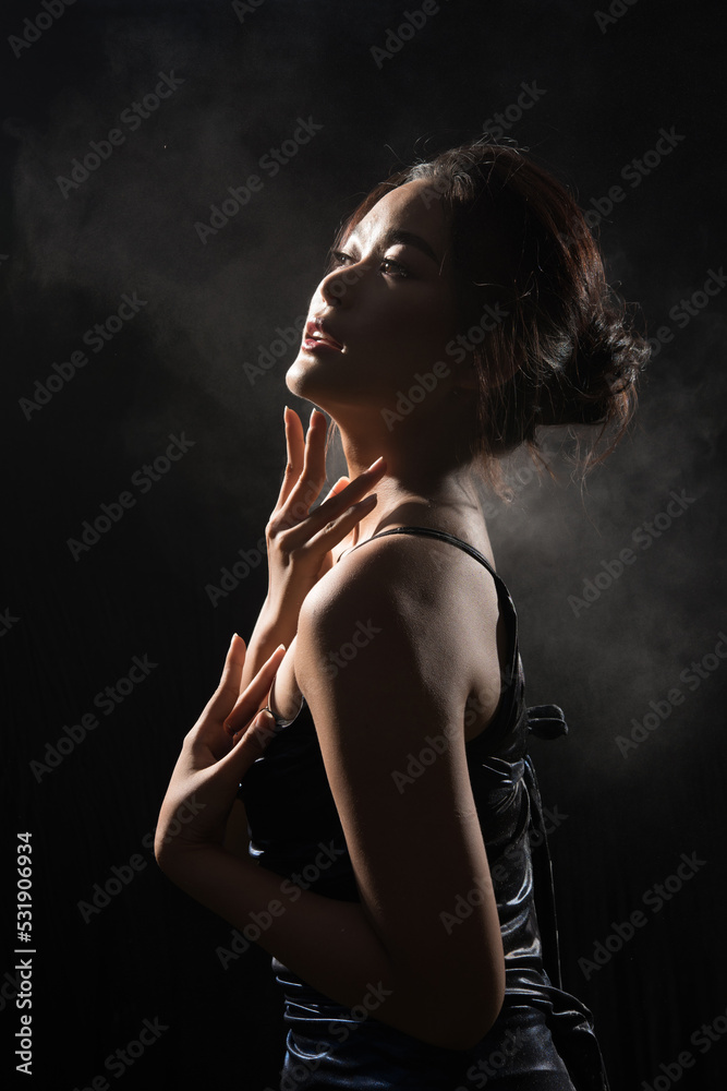 Naklejka premium Portrait of Asian Woman in shadow low light key with back backlit, side rear view of female turn twist body for healthy shape fashion style. black background smoke exposure
