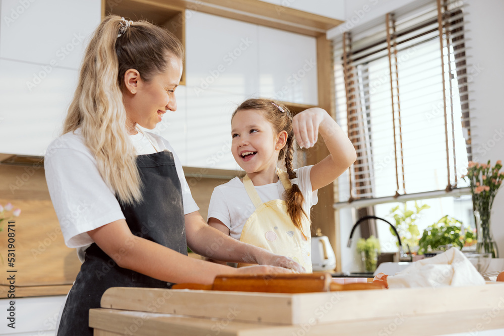 Young adorable mother teaching kid kneading dough preparing domestic cookies biscuits apple pie surprise for family. Girls enjoying process laughing having fun together at weekend baking cooking. Young adorable mother teaching kid kneading dough preparing domestic cookies biscuits apple pie surprise for family. Girls enjoying process laughing having fun together at weekend baking cooking.