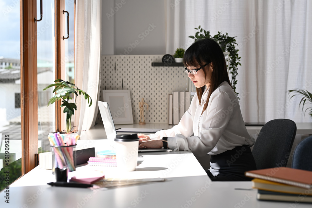Side view asian female employee using laptop computer, working on marketing project in bright office Side view asian female employee using laptop computer, working on marketing project in bright office