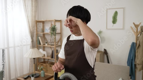 slow motion of a tired asian man wearing household apron is wiping sweat on his forehead while doing house chores with a vacuum cleaner in the living room at home