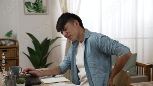 slow motion of tired asian business man sitting at desk rubbing massaging his painful lower back with a grimacing face while working from home in the living room