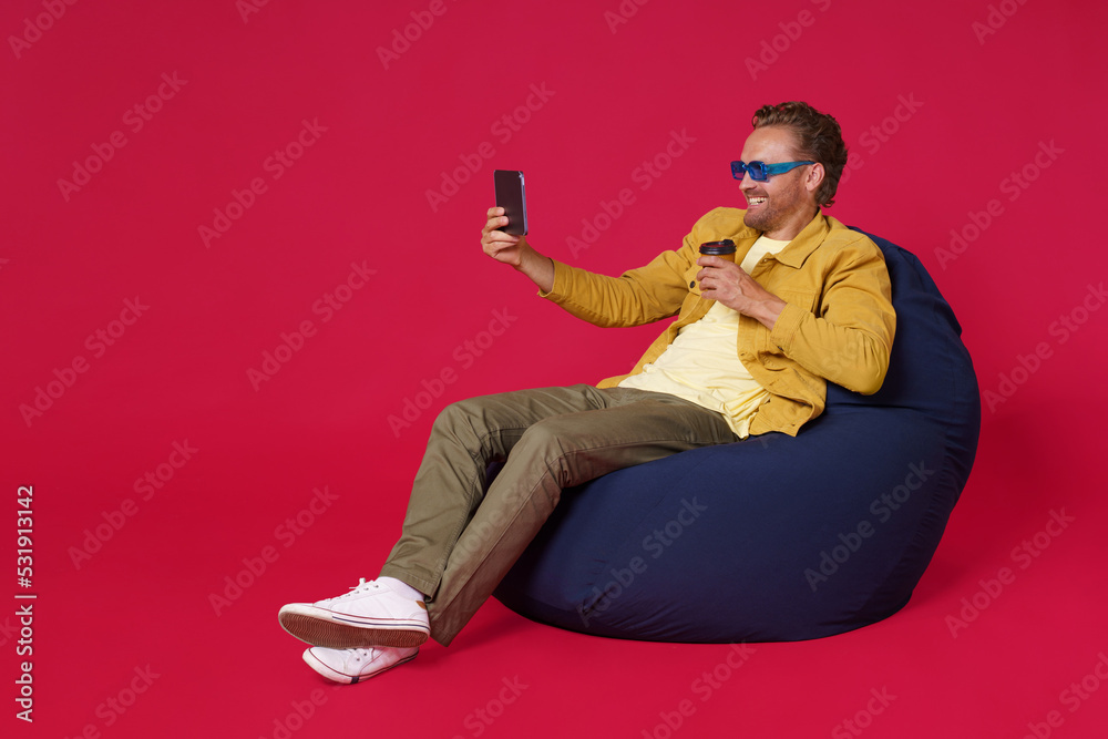Happy handsome young man 30s make selfie using digital tablet drinking coffee from paper cup wearing casual denim outfit sitting on bag chair isolated on red background. Coffee shop business concept Happy handsome young man 30s make selfie using digital tablet drinking coffee from paper cup wearing casual denim outfit sitting on bag chair isolated on red background. Coffee shop business concept