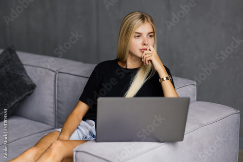 Pensive young woman using laptop while sitting on a couch at home