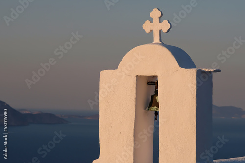A cross and a bell on the top of an orthodox church in Ios Greece while the sun is setting in the background
