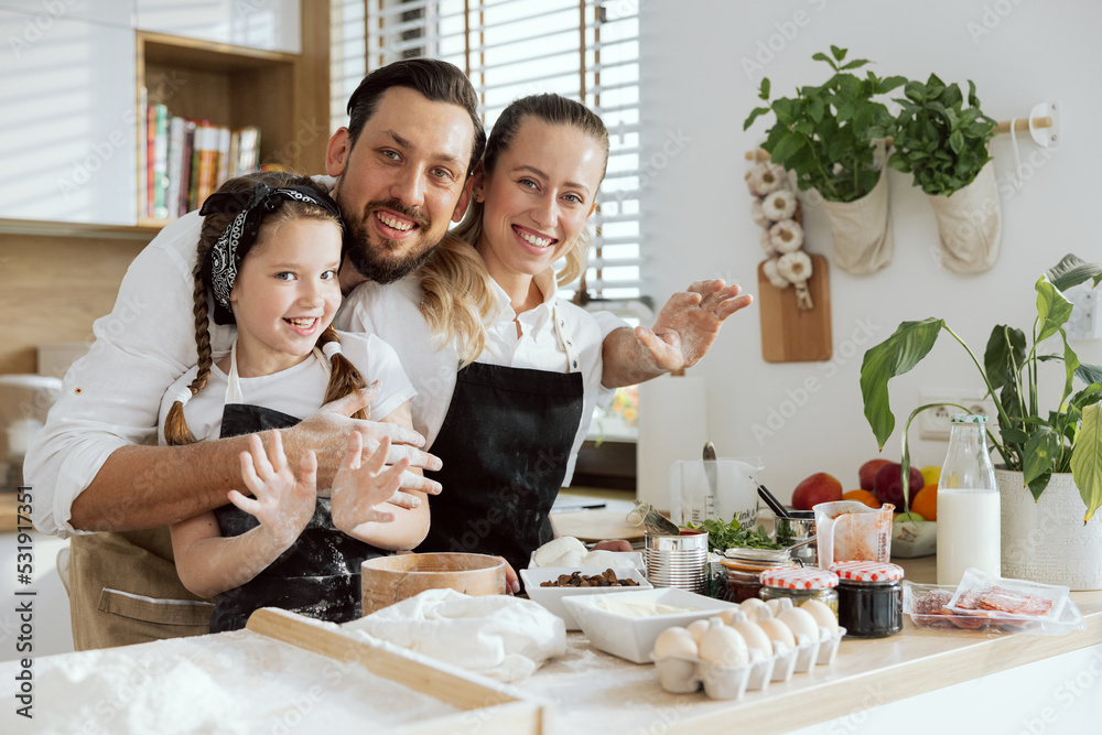 Adorable family wearing aprons looking at camera smiling spending time together on modern kitchen. Tasty ingredients on wooden surface eggs flour mushrooms prepared for dressing homemade pizza.