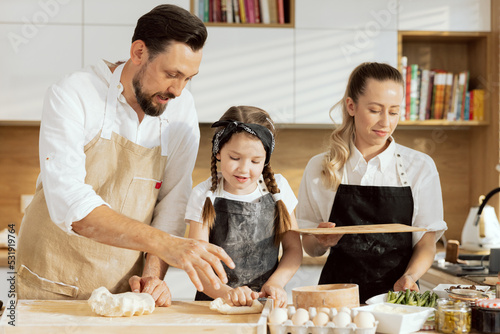 Happy family in modern kitchen. Parents teaching daughter kneading homemade dough for baking domestic pizza in moderm light kitchen. Delighted beautiful mother in apron helping holding wooden desk.