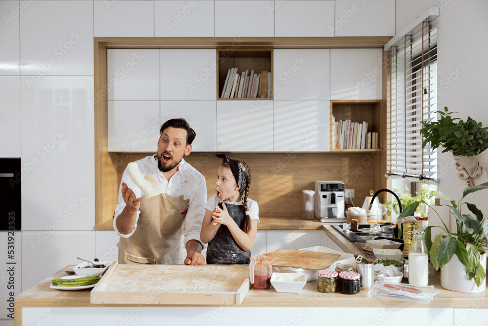 Excited girl kid daughter with surprising emotions clapping in hands looking how father dad with beared throwing up dough for pizza. Pizza ingredients on wooden table. Girl having fun with dad. Excited girl kid daughter with surprising emotions clapping in hands looking how father dad with beared throwing up dough for pizza. Pizza ingredients on wooden table. Girl having fun with dad.