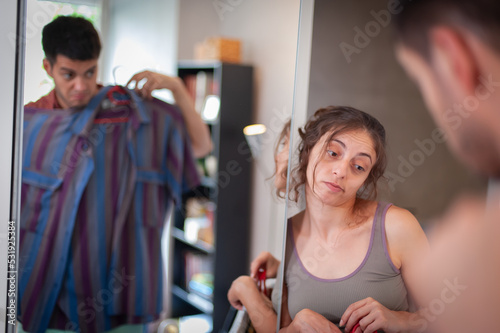 A charming young man tries on clothes. He is standing in front of the bedroom mirror, next to him is a woman helping him choose a shirt.