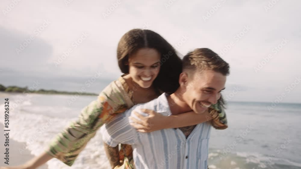 Happy young caucasian man carrying girlfriend on back while laughing at the beach Happy young caucasian man carrying girlfriend on back while laughing at the beach