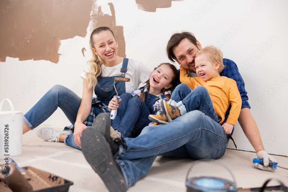 Smiling and happy family of four sitting on the floor. Around them are repairs. They are happy and discuss whether everyone likes the new brown wall color.