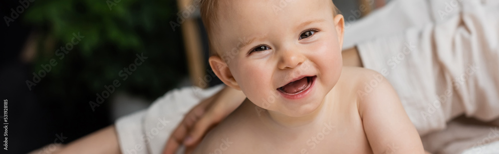 mother near happy infant daughter looking at camera in bedroom, banner.
