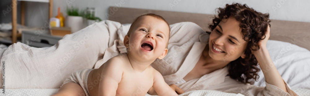 happy and curly mother smiling while looking at excited infant daughter on bed, banner. happy and curly mother smiling while looking at excited infant daughter on bed, banner.