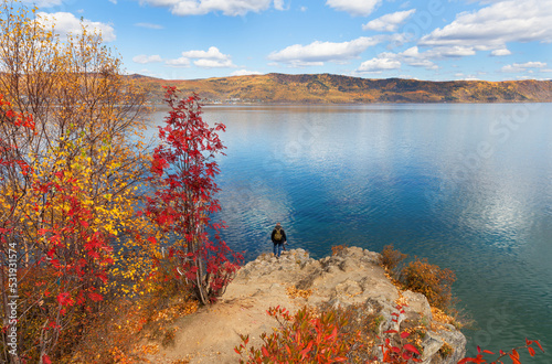Autumn landscape of Baikal Lake. A tourist with a camera travels along Cape Shamansky. Yellowed birches and red rowan trees in a forest on a rocky shore on a sunny September day. Natural background