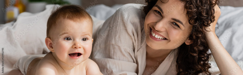 happy woman with curly hair smiling while looking at camera near excited infant baby, banner. happy woman with curly hair smiling while looking at camera near excited infant baby, banner.