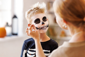 Festive makeup for Halloween. Woman doing skeleton make-up for boy in costume while preparing holiday