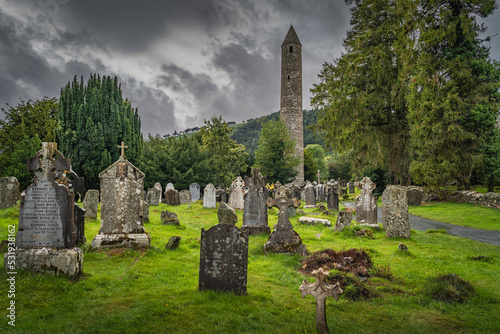 Medieval tower and ancient graves with Celtic crosses in Glendalough Cemetery. Moody storm sky in background, Wicklow, Ireland