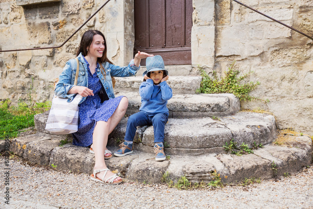 Fototapeta premium Mother and little handsome baby boy sitting on ancient stone stairs and playing outdoor with straw hat in old town