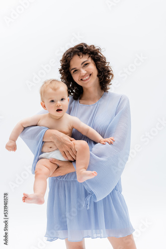 Positive curly woman in dress holding baby in panties isolated on white.