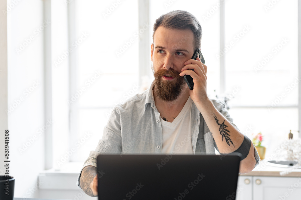 Young bearded man working from home with laptop while talking on the phone Young bearded man working from home with laptop while talking on the phone