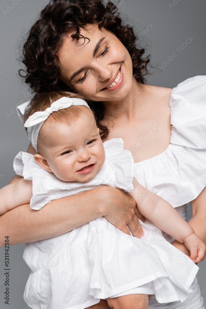 Joyful woman hugging smiling baby daughter in dress isolated on grey. Joyful woman hugging smiling baby daughter in dress isolated on grey.