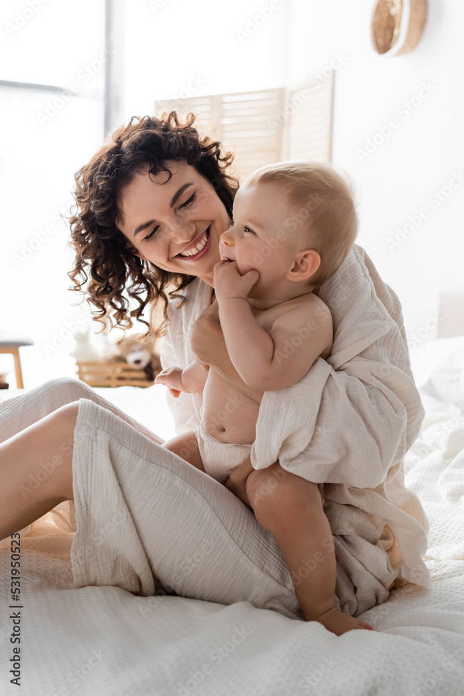 cheerful woman in loungewear holding in arms baby daughter and sitting on bed. cheerful woman in loungewear holding in arms baby daughter and sitting on bed.