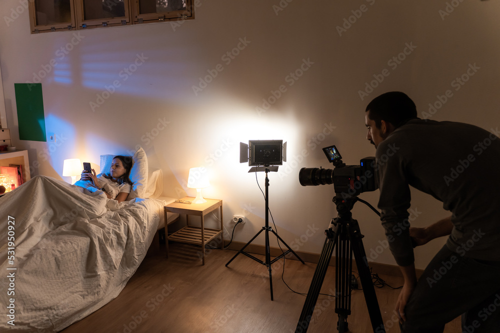 Film bedroom set with attractive model in the bed and camera operator with lights. Behind the scenes.