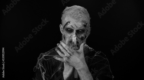 Zombie man with makeup with fake wounds scars and white contact lenses spells conjures over a candle, trying to scare in dark studio room. Sinister dead guy. Halloween, filming, staging concept