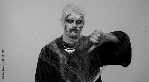Sinister man in carnival costume of Halloween crazy zombie with bloody wounded scars face showing thumbs down sign gesture, expressing discontent, disapproval, dissatisfied, dislike. Cosplay undead