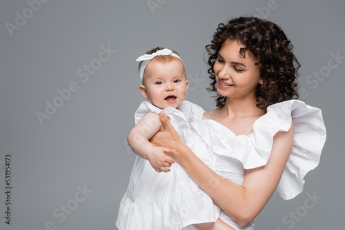 Curly woman holding cheerful baby girl in dress isolated on grey.