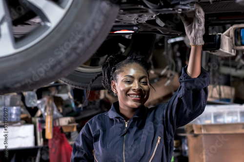 Smiling African black car mechanic woman working underneath car in auto repair shop, Car maintenance concept