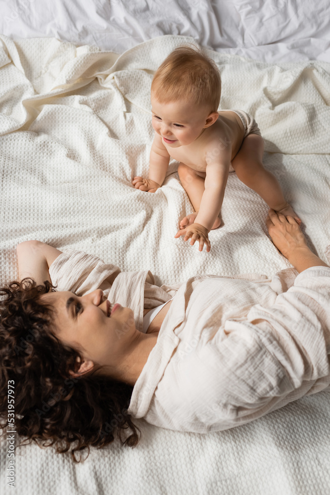 top view of curly mother smiling while looking at cheerful infant daughter on bed.