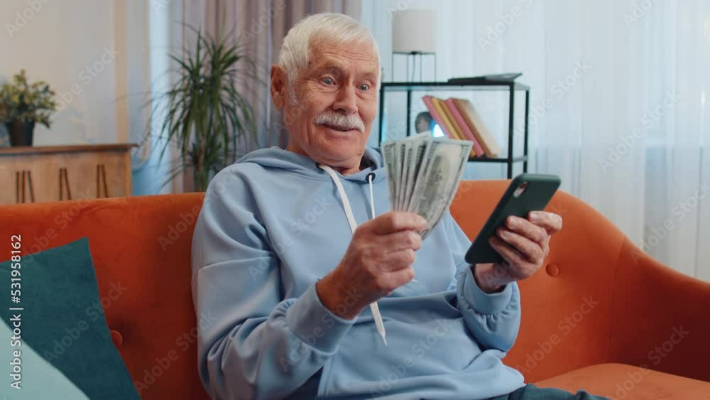 Planning family budget. Smiling senior old grandfather counting money cash, use smartphone calculate domestic bills at home. Joyful man satisfied of income and saves money for planned vacation, gifts Planning family budget. Smiling senior old grandfather counting money cash, use smartphone calculate domestic bills at home. Joyful man satisfied of income and saves money for planned vacation, gifts