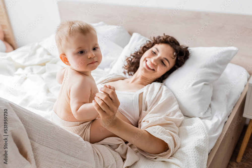 happy woman in loungewear lying on bed and holding hand of infant daughter. happy woman in loungewear lying on bed and holding hand of infant daughter.