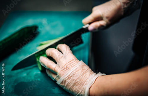 woman chef hands in disposable gloves slicing cucumber on cutting board.